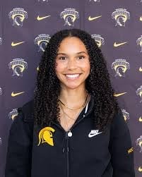 Woman with long black curly hair wearing black Nike jacket with yellow and black athletic logo, standing in front of branded repeating logo backdrop