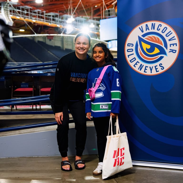 Two people posing together inside a Vancouver Canucks arena, with one wearing a Canucks jersey and holding a merchandise bag