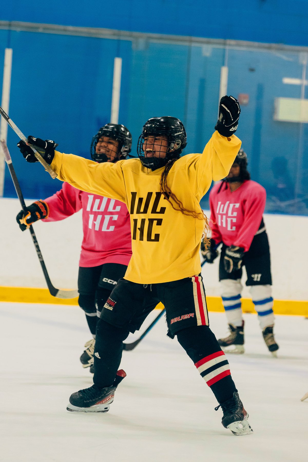 Young hockey player in yellow jersey celebrates with arms raised on ice rink, teammates in pink jerseys nearby