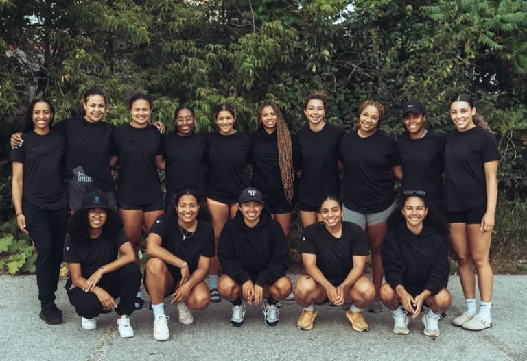 Group of eleven women in matching black t-shirts posing together outdoors with green foliage in background