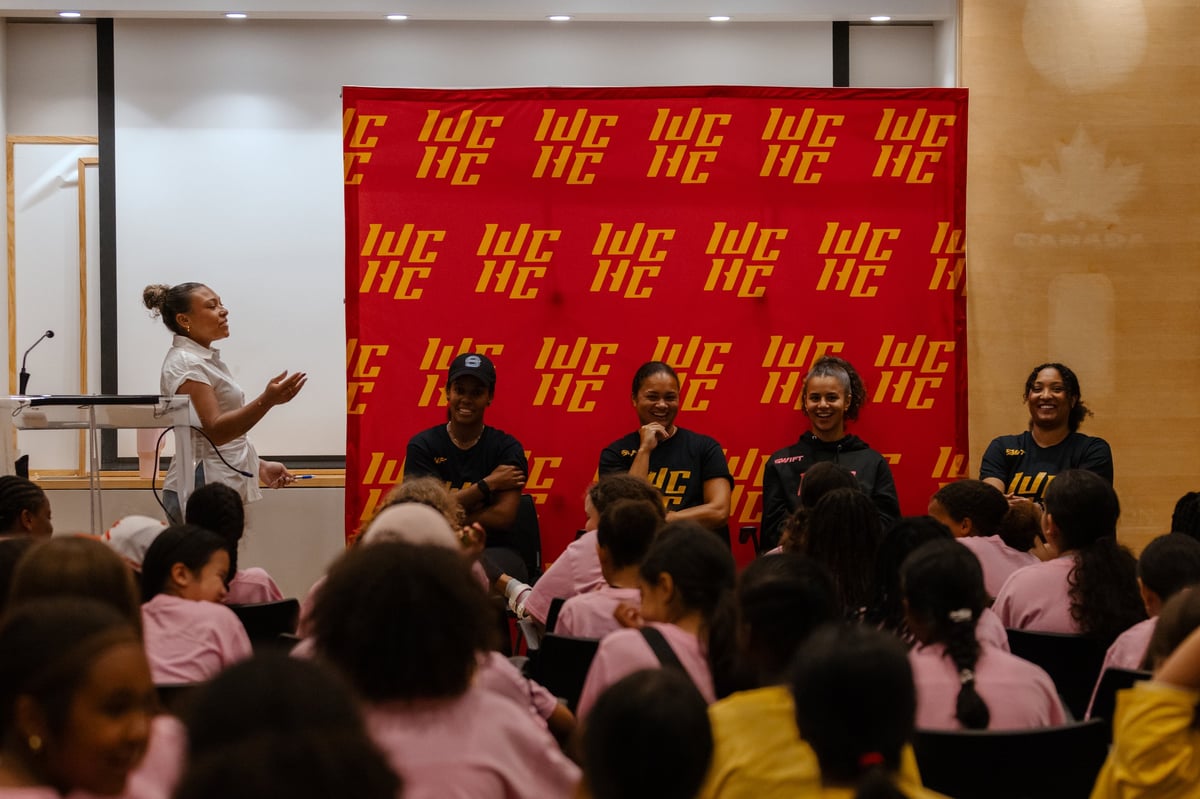 A speaker gestures while addressing an audience seated on the floor in front of a red and gold branded backdrop