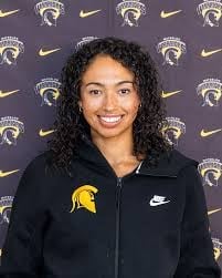 Young woman with curly black hair wearing a black Nike zip-up jacket with a yellow logo, smiling in front of a branded backdrop