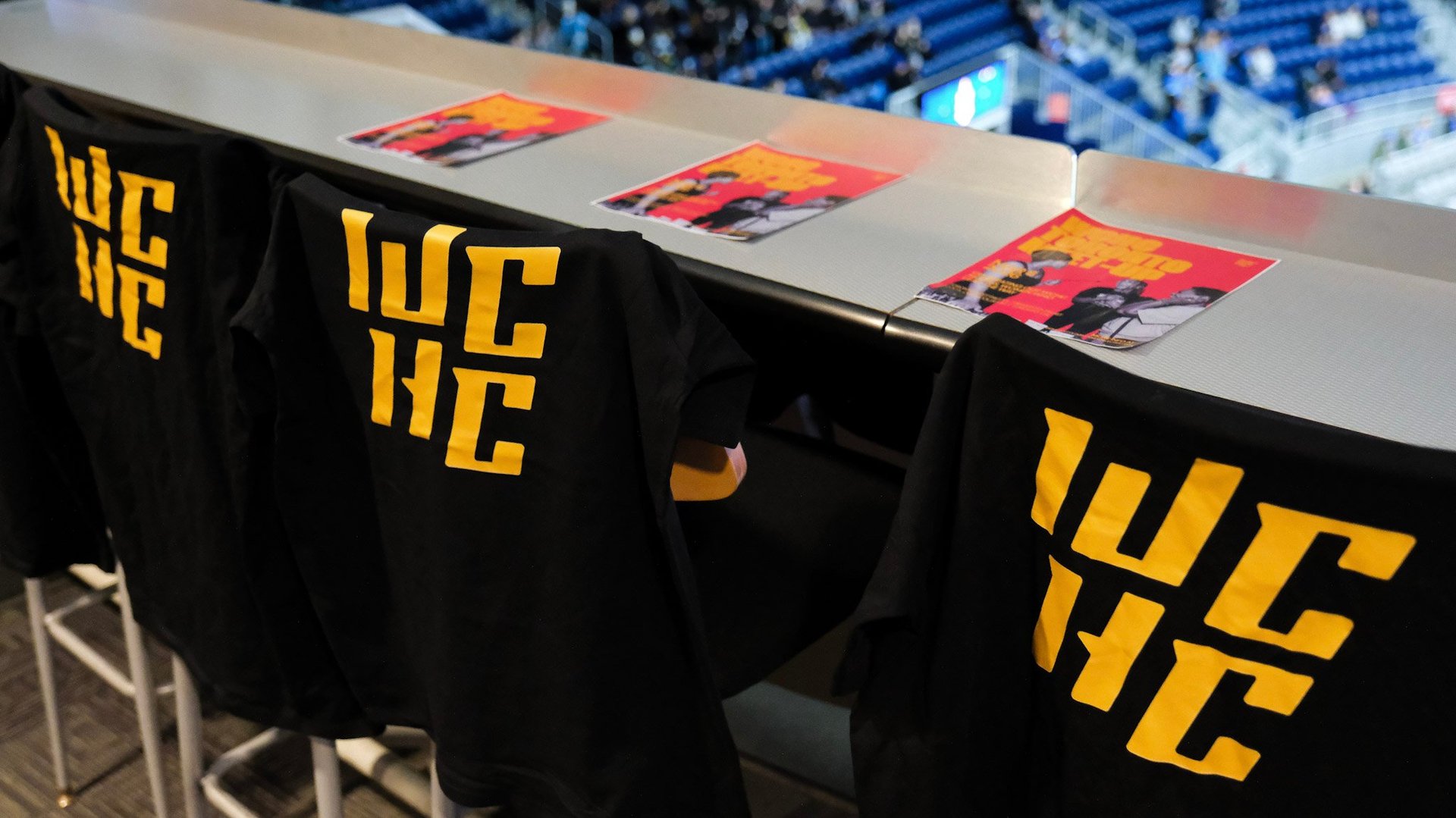 Black branded bags with yellow logo displayed on a table at a stadium event with blue seating visible