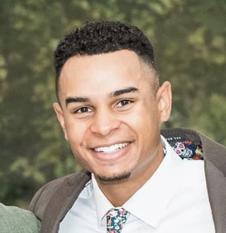 Denzel Radford smiling, wearing a navy blazer over a white top with a green blurred background