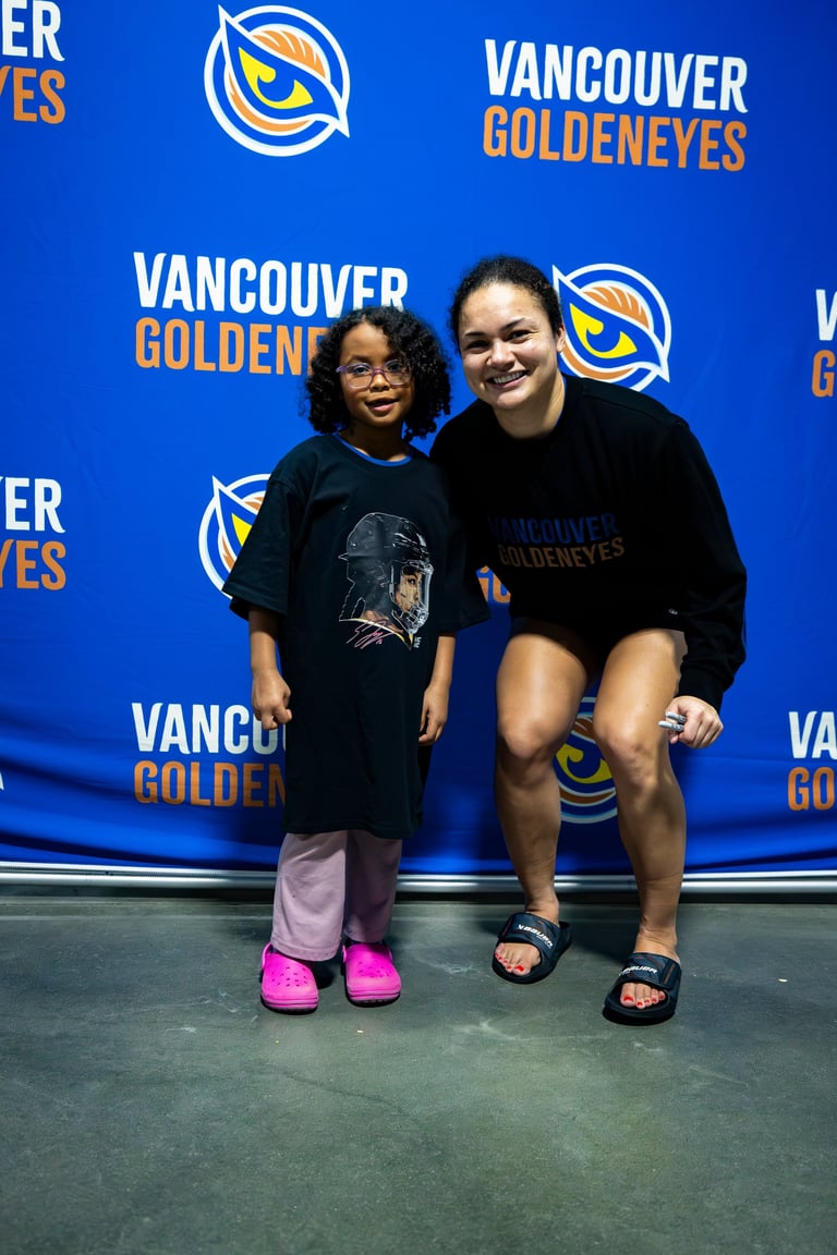 Two people posing in front of a blue Vancouver Goldeneyes branded backdrop at what appears to be a promotional event or photo opportunity