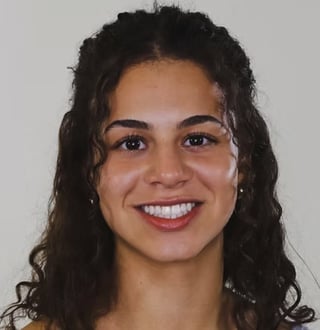 Jade Iginla smiling with long dark wavy hair and small earrings against a light gray background
