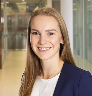 Stephanie Sucharda smiling with long brown hair wearing a dark blazer over a white shirt in a modern office hallway