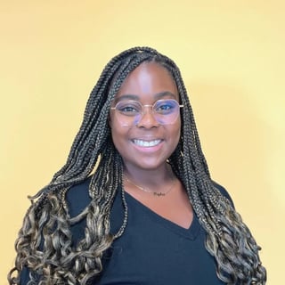 Kyla Mitchell smiling with long braids and glasses, wearing a black top against a pale yellow background