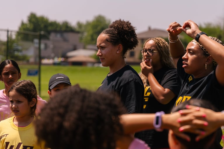 Group of young athletes in yellow and black team shirts gathered on a sunny field with green grass and buildings visible in the background
