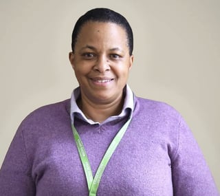 Kim Borden Penney smiling, wearing a purple sweater with a green lanyard against a beige background